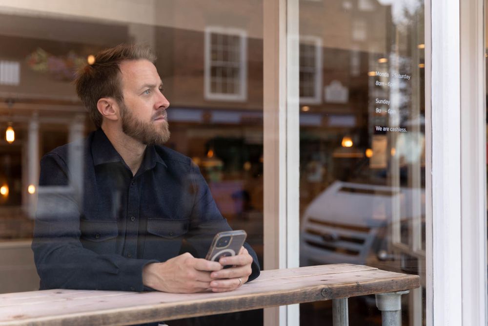 Personal branding photograph taken through a window of a life coach holding his mobile phone, looking out into the distance