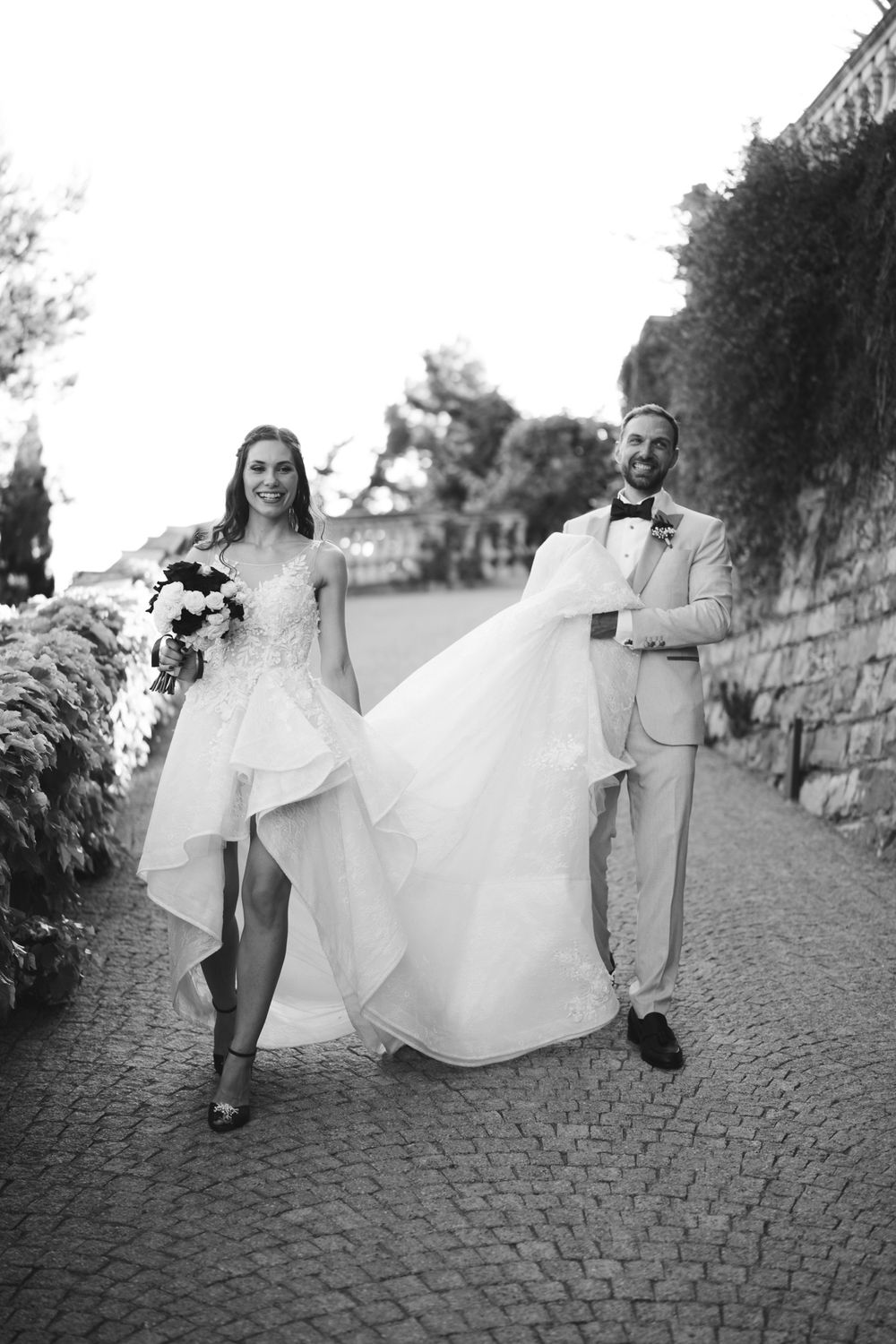 A smiling bride and groom walk through the garden of Villa Casa Rossa in Imperia during a summer wedding.