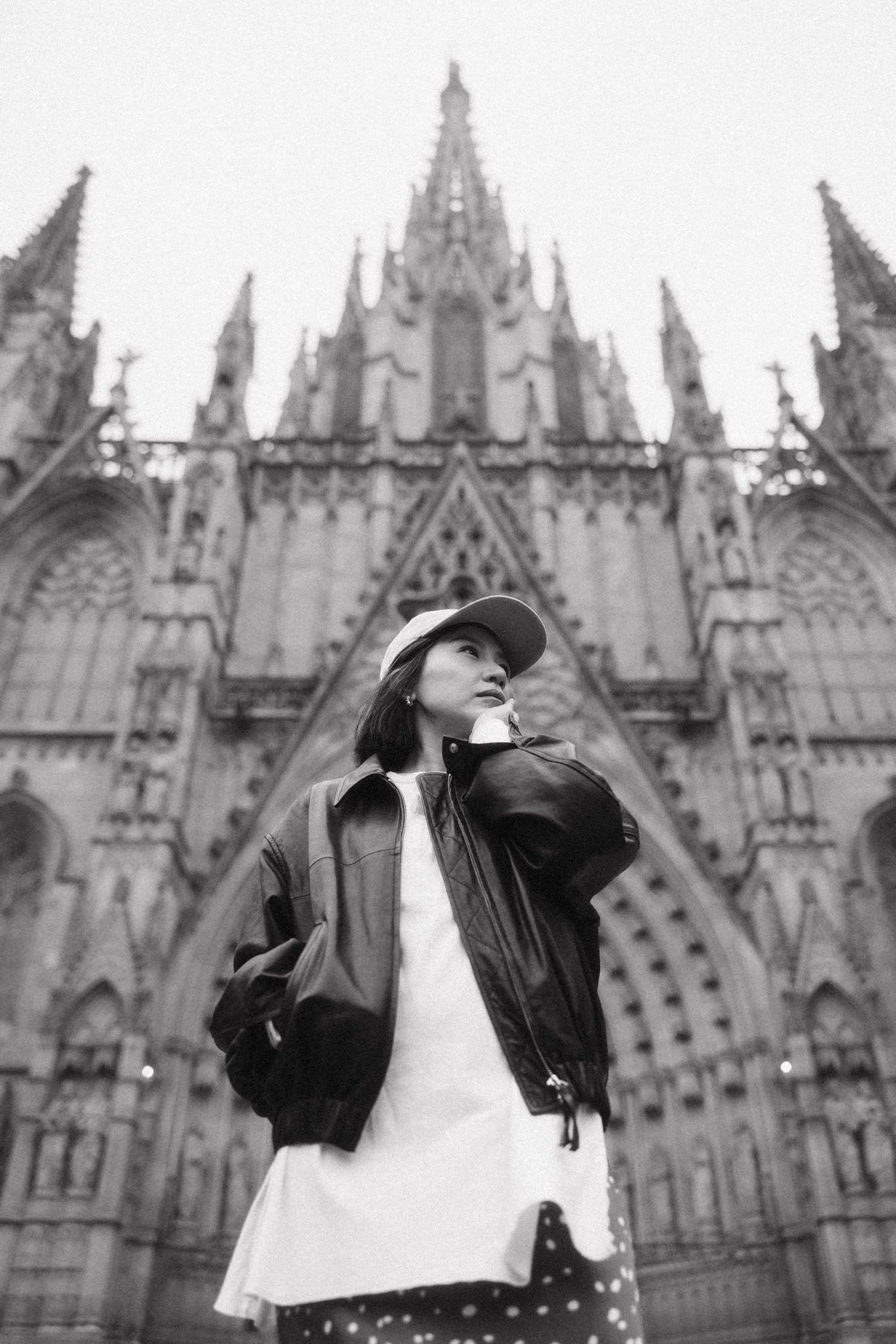Young woman in leather jacket and floral skirt standing before Gothic cathedral spires in Barcelona, Spain.