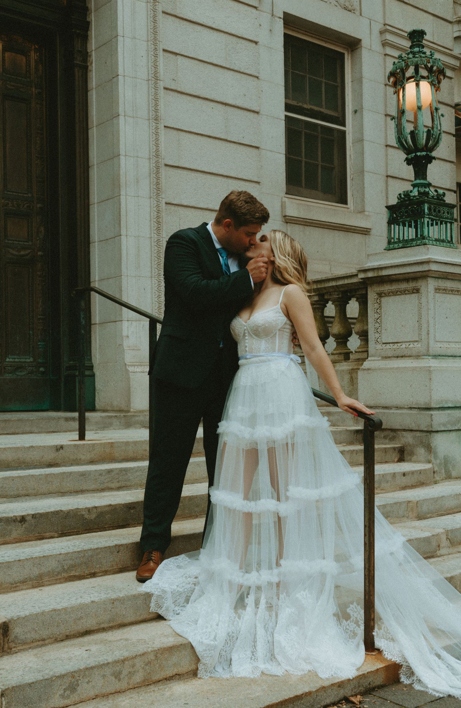 Bride and groom share a romantic kiss on stone steps outside an elegant building, bride wearing a tiered tulle gown.