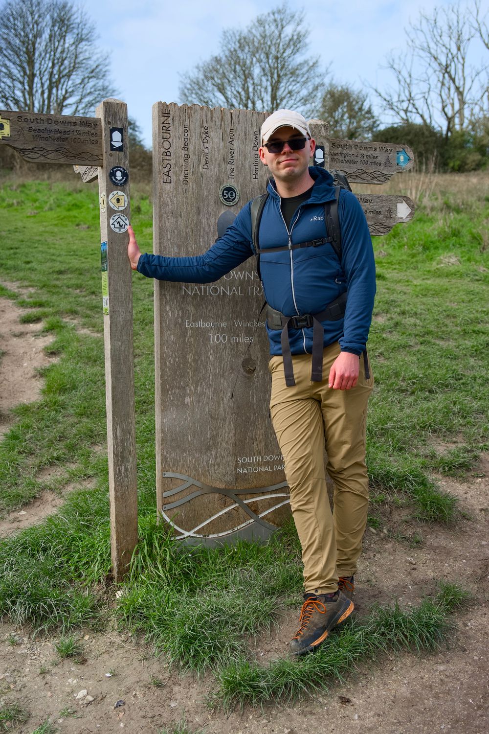 Hiker in blue jacket and tan pants poses beside a wooden trail sign in a national park on a cloudy day.