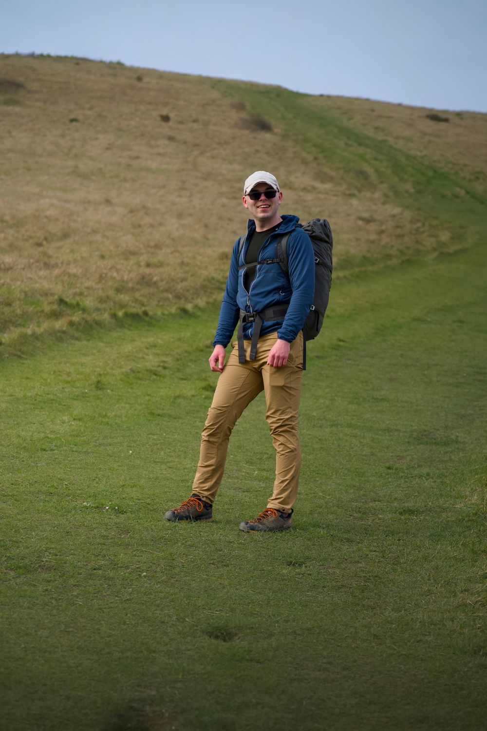 A male hiker wearing a blue jacket, tan pants, and backpack stands on a green hillside trail on a cloudy day.