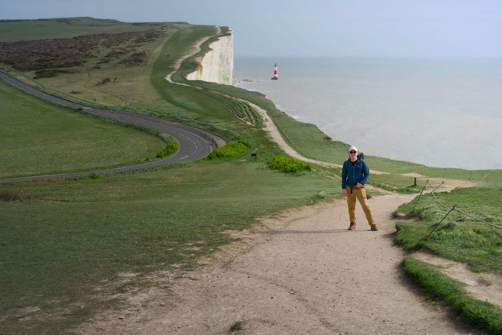 Person standing on coastal cliff path with white chalk cliffs, lighthouse, and green hills overlooking the sea.