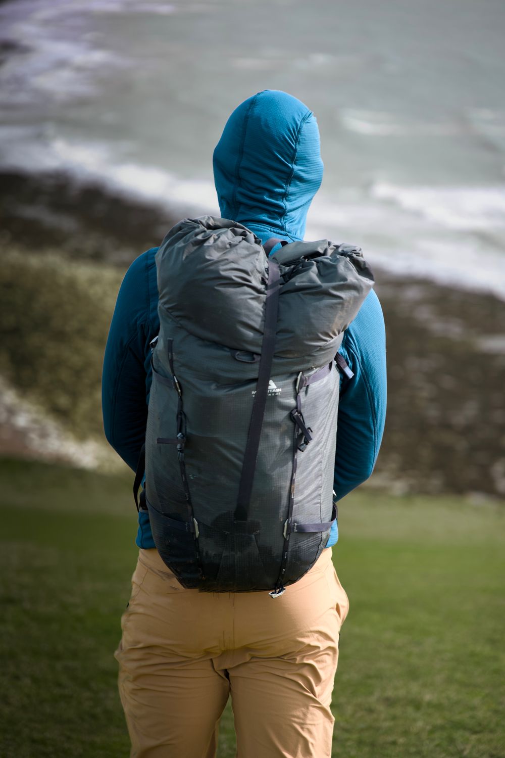 Hiker in blue hooded jacket with gray backpack stands facing a rocky shoreline with waves in the background.