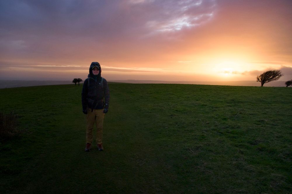 Person standing on green hillside at dramatic orange sunset with windswept trees and colorful cloudy sky.