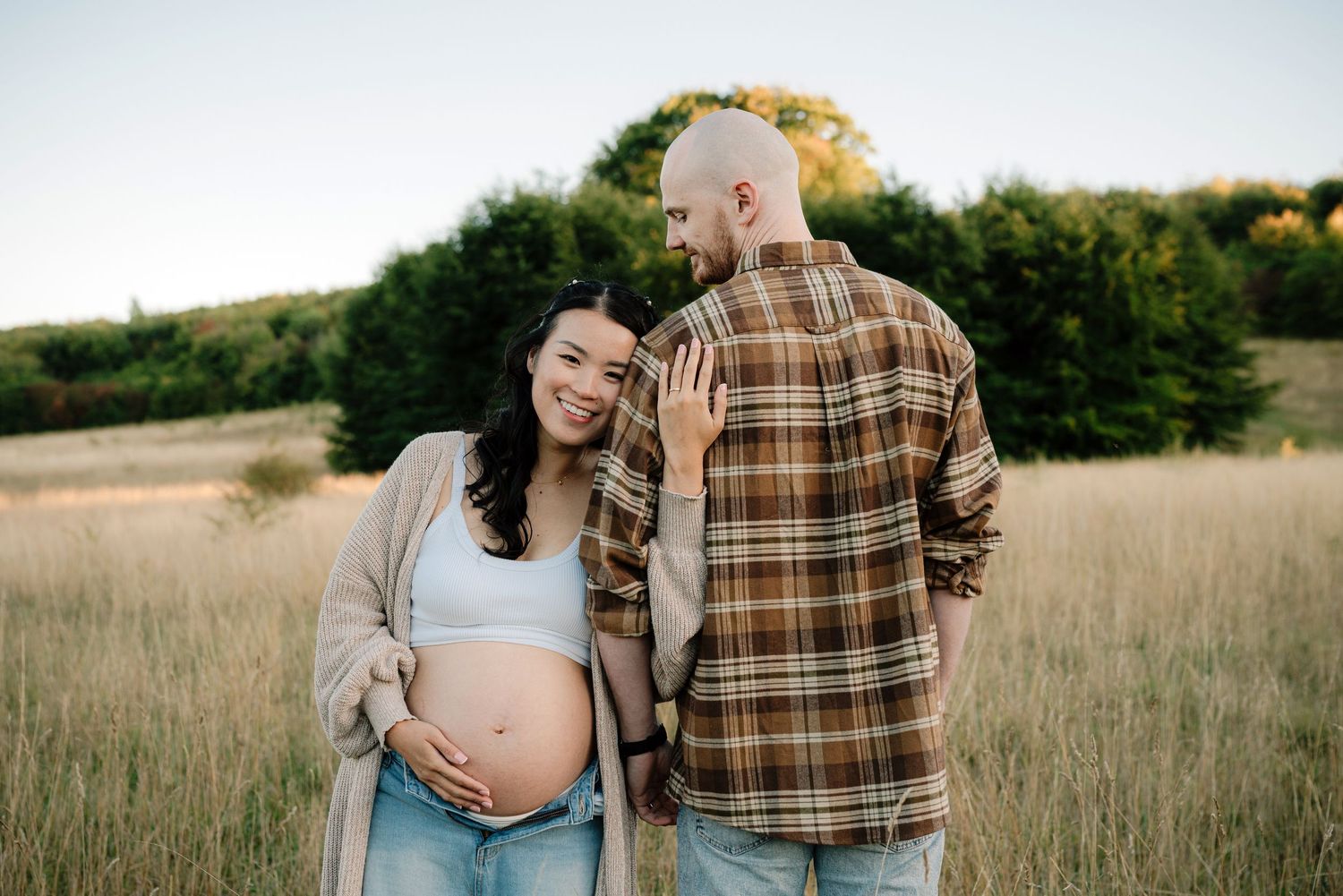 A pregnant woman and her partner stand in a field, embracing each other and enjoying the natural surroundings.