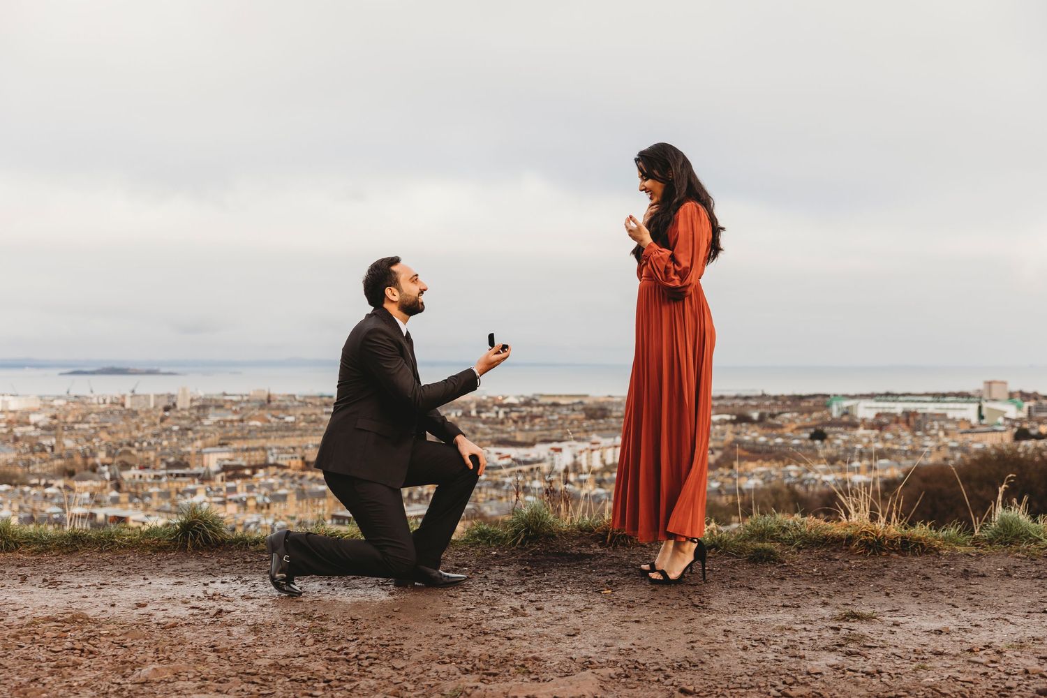 A romantic proposal scene overlooking a cityscape, with a person kneeling before another in a flowing red dress.
