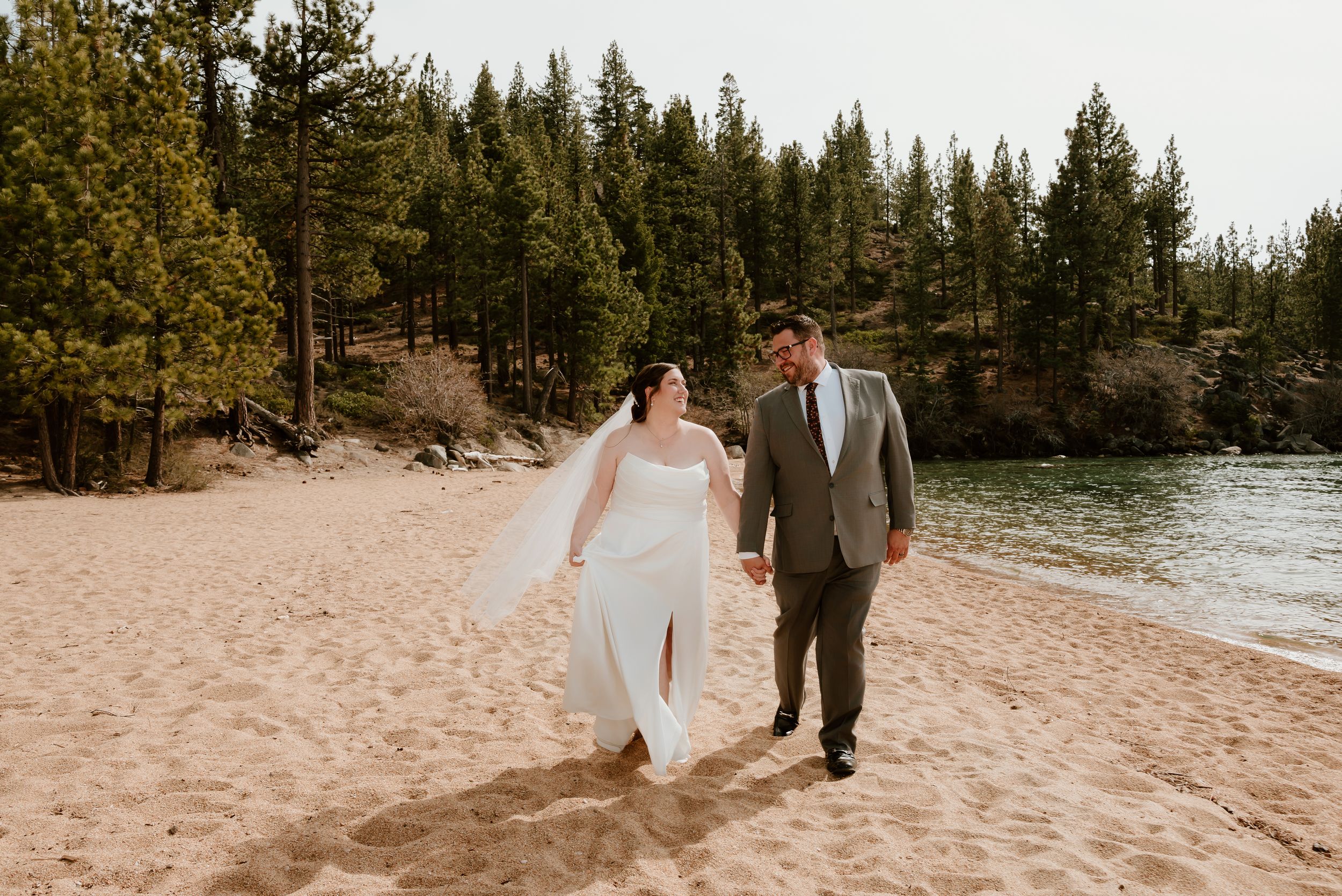 Couple in wedding attire walking along a sandy beach with evergreen trees and lake in the background.