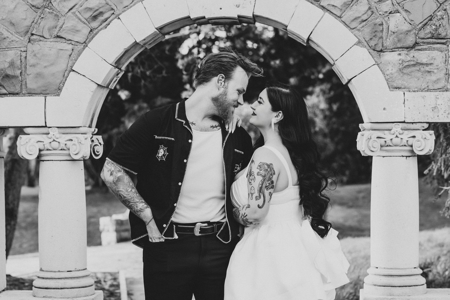 Black and white photo of tattooed bride and groom sharing romantic moment under stone arch at outdoor wedding ceremony.