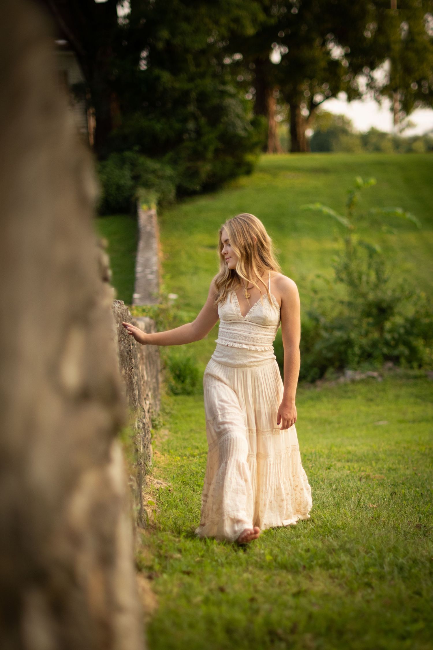 A person in a flowing cream-colored dress walks through a lush green garden at sunset.