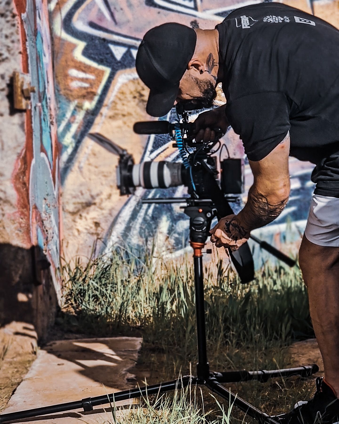 Videographer in black clothing operates professional camera equipment near graffiti wall and tall grass in outdoor setting.