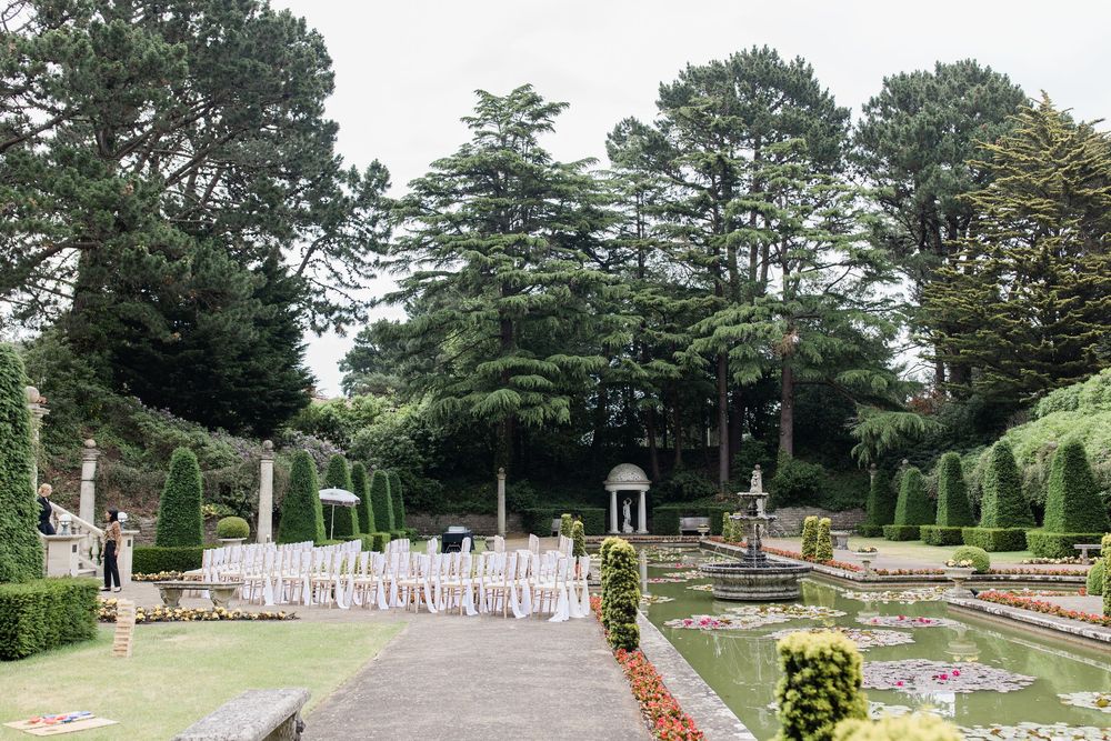 White chairs arranged in rows for an outdoor wedding ceremony in a formal garden with tall hedges and trees at The Italian Villa in Dorset