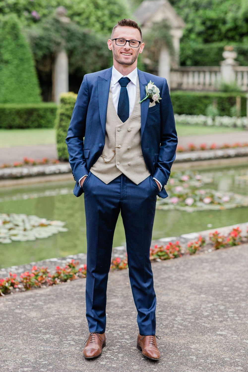 Groom in navy suit and tan vest poses in formal garden with blooming flowers at Italian Villa Wedding