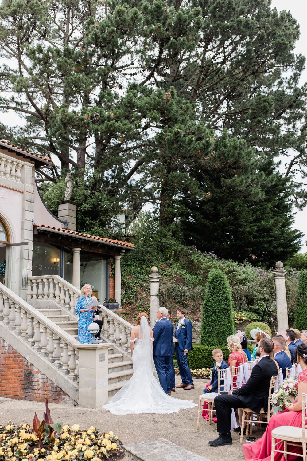Wedding ceremony taking place at an elegant outdoor venue with stone stairs and columns surrounded by pine trees by Maddie Waters