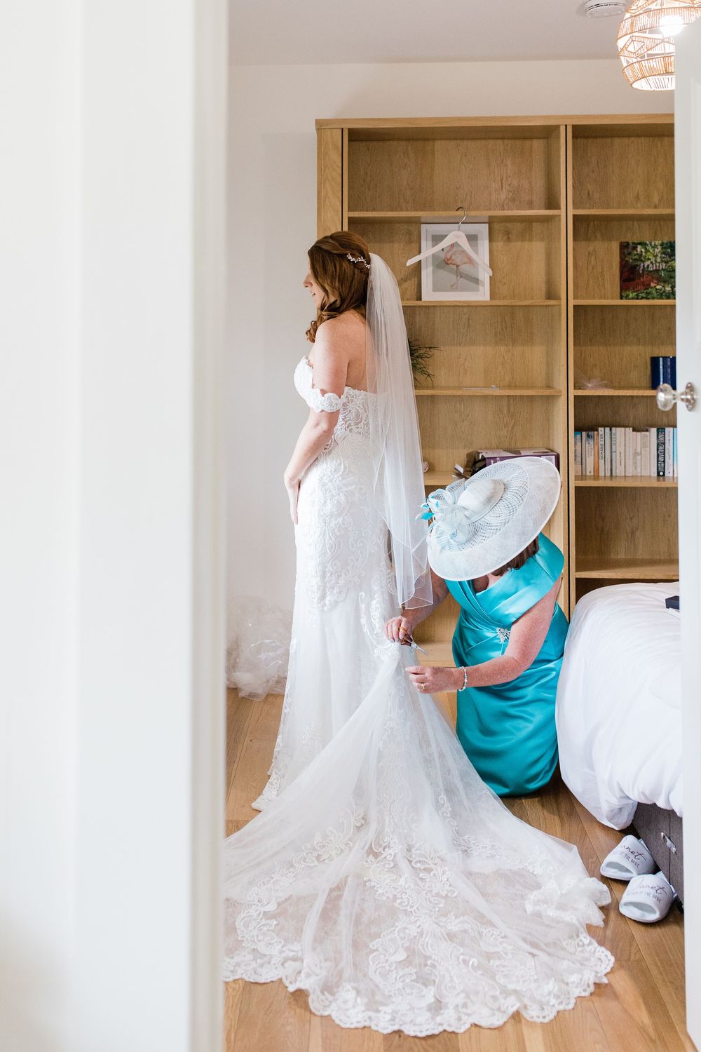A bride in a lace wedding dress with a flowing train gets final adjustments from an attendant in turquoise by Maddie Waters Photographer