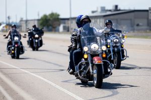 A group of motorcyclists riding in formation on a road with buildings in the background.