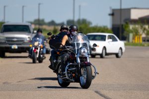 Motorcyclists riding in a group on a road with cars and buildings in the background.