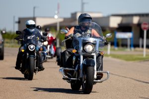 Motorcyclists riding in formation on a road with buildings and a clear blue sky visible.