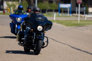 Two motorcyclists riding large touring bikes on a road with buildings in the background.