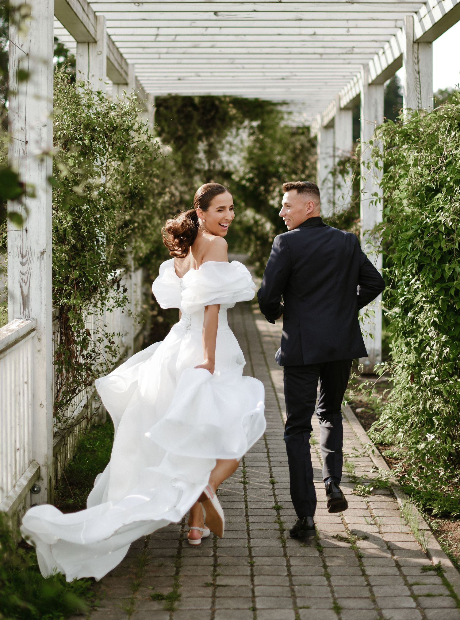 Bride and groom walking through a white pergola garden path with flowing wedding dress.