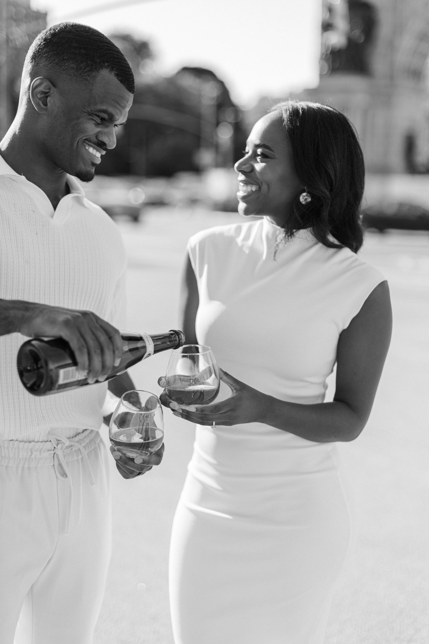 A couple in white outfits share champagne together outdoors.