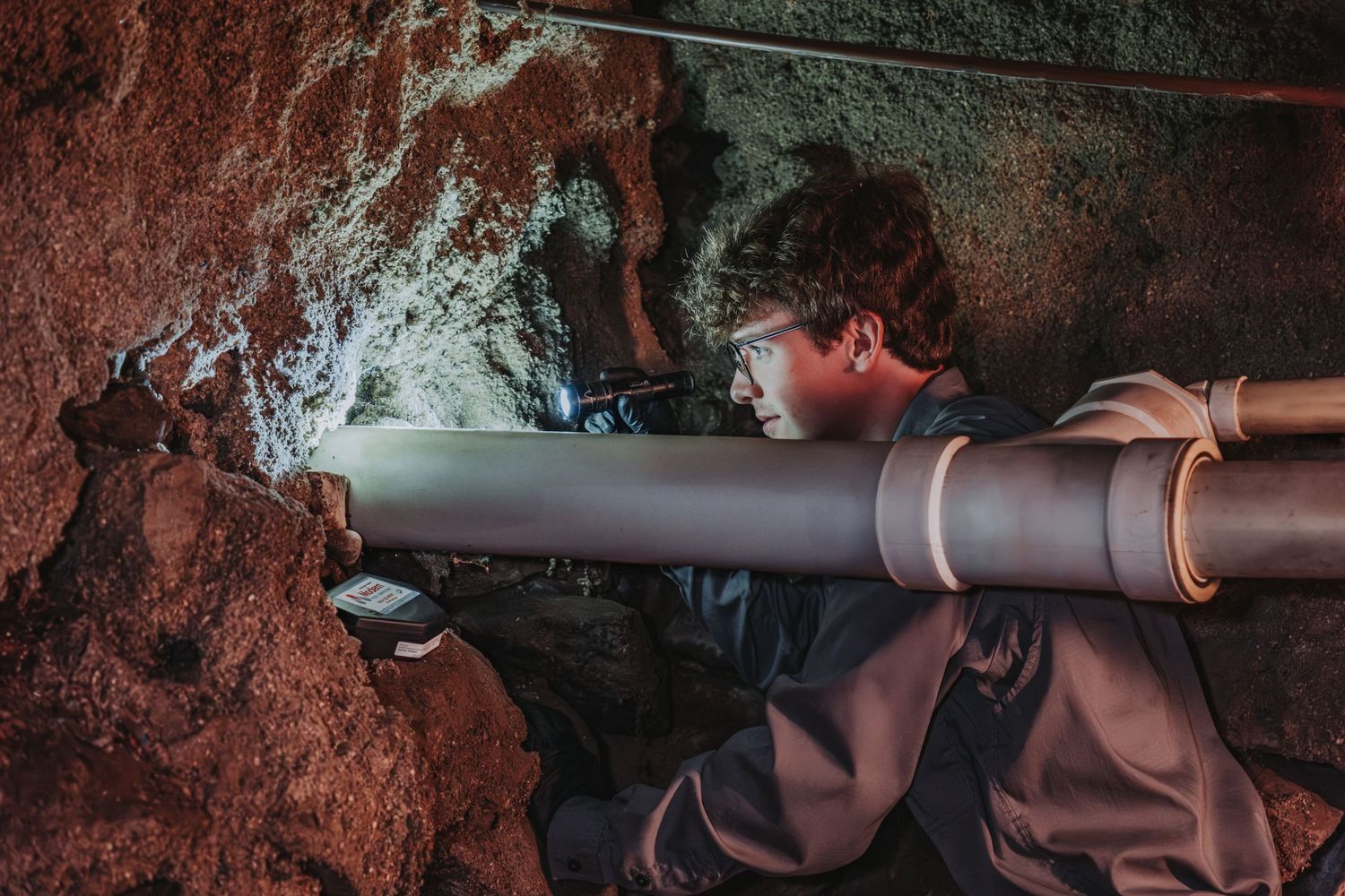 A Modern Pest Control Employee is inspecting a large industrial pipe system against a textured rock wall in dim lighting in a  Maine home.