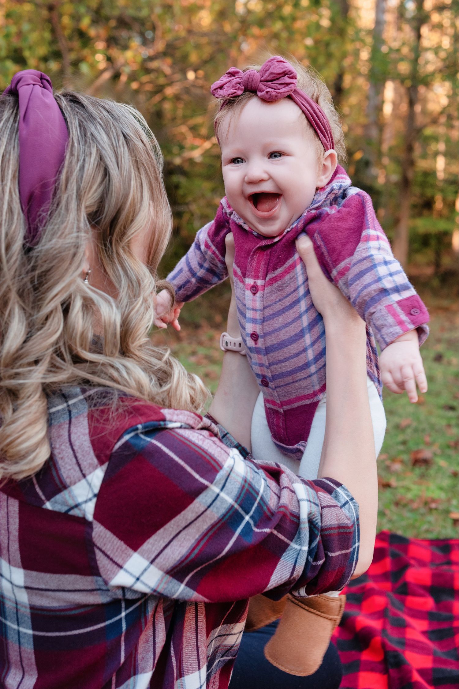 A joyful baby in a purple outfit being lifted up outdoors on a plaid blanket.