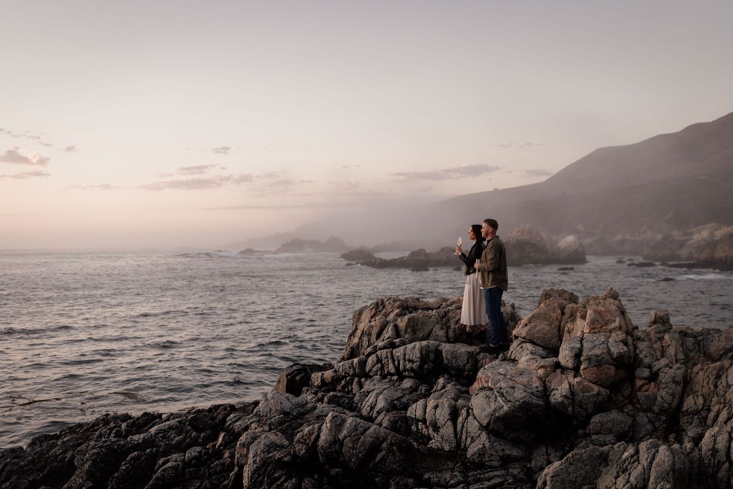 Romantic sunset silhouette on rocky coastal overlook with mountains and ocean in background.
