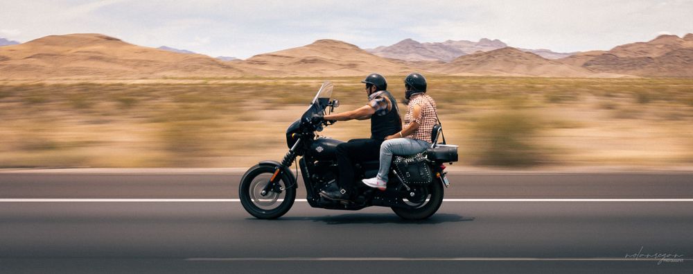 A motorcycle rides through a desert landscape with mountains in the background.