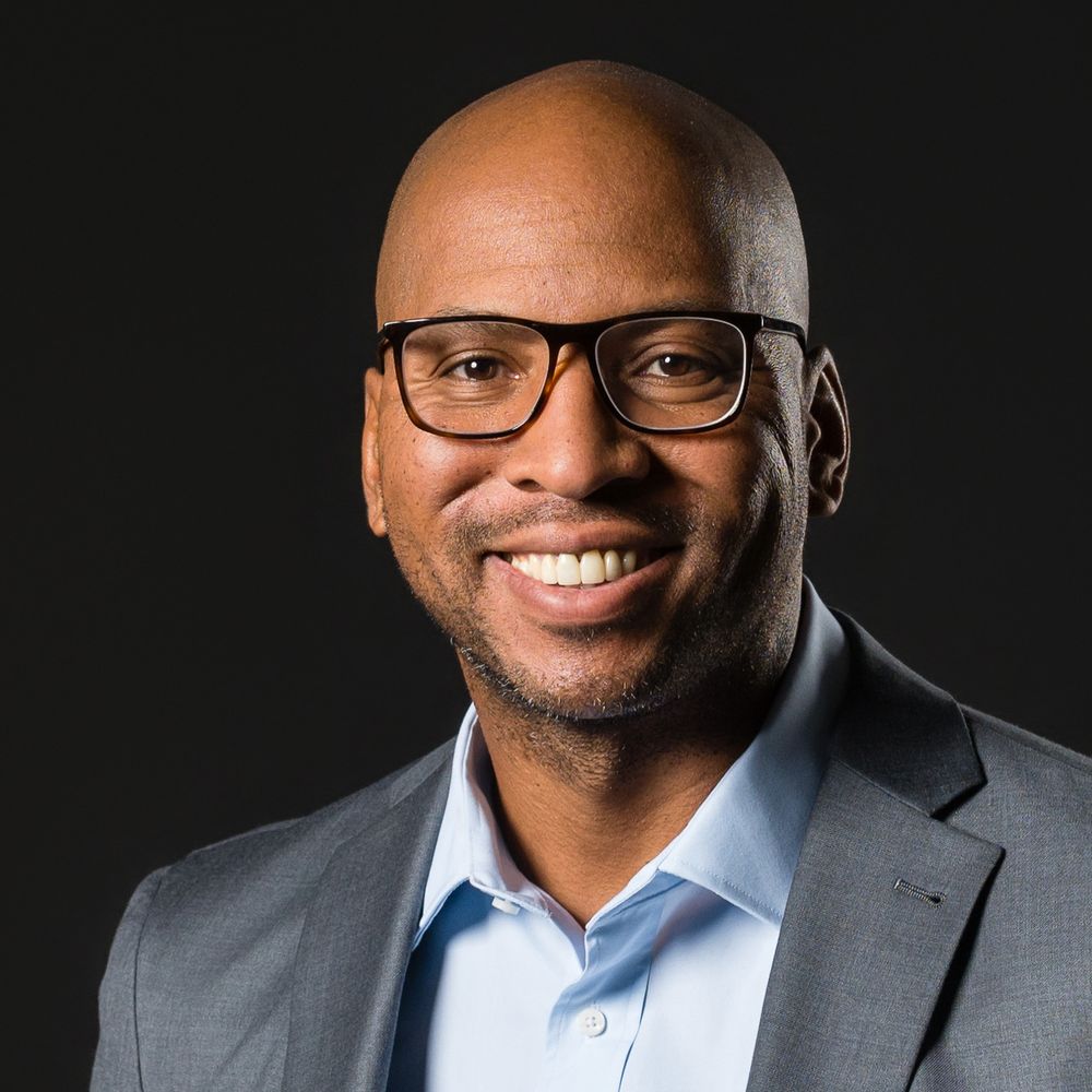 Professional headshot of a smiling person wearing glasses, a blue shirt and grey blazer against a dark background.