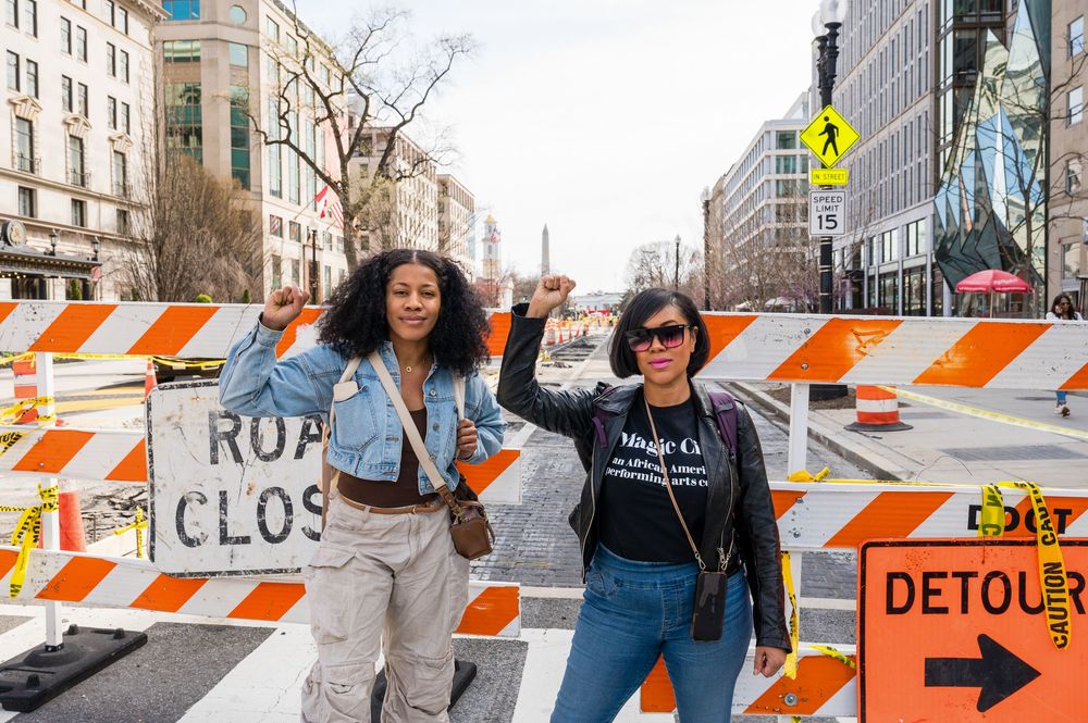 Construction detour signs and barriers line a city street with orange and white striped barricades.