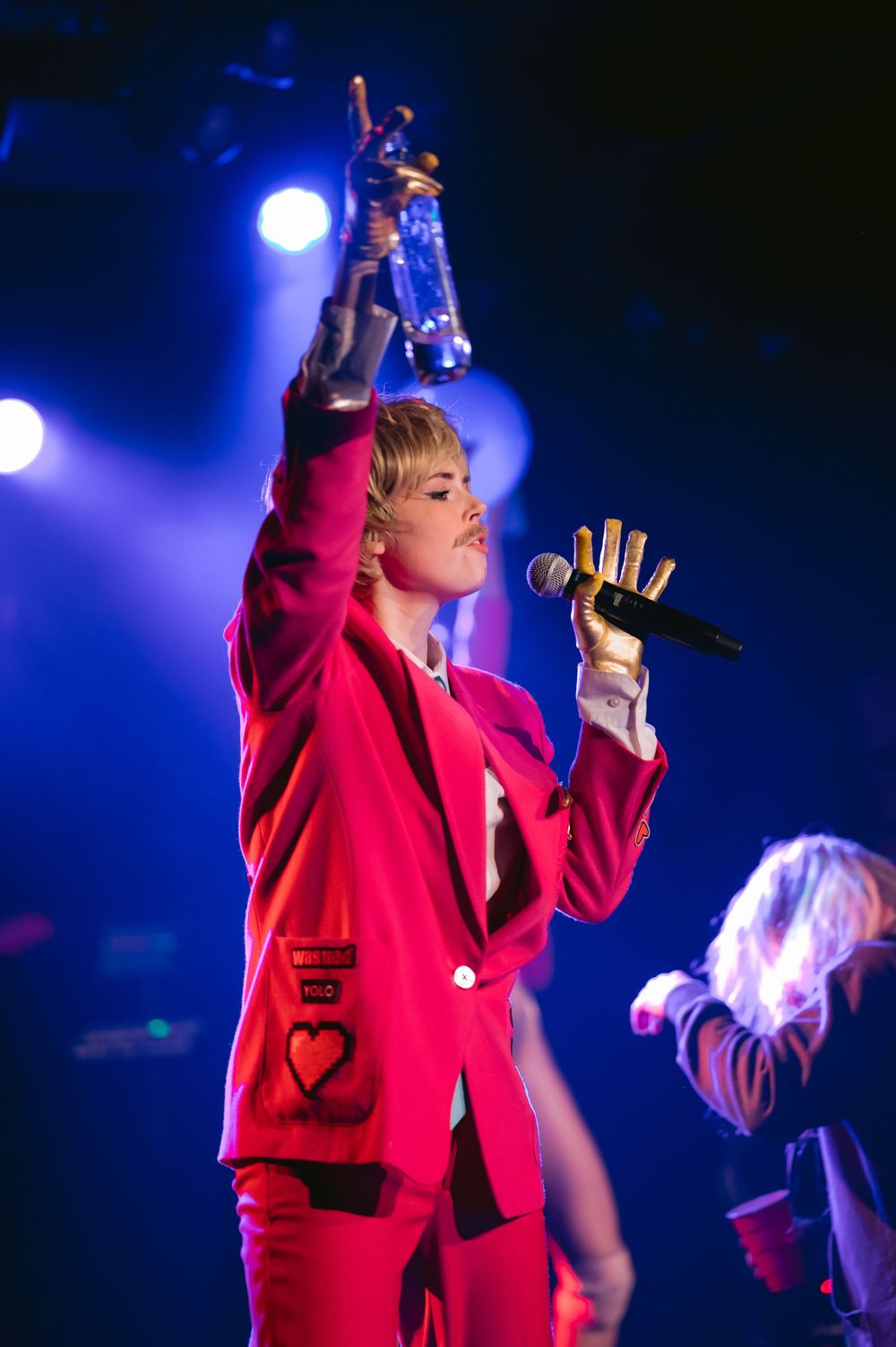 A performer in a bright red suit jacket sings passionately on stage under dramatic blue lighting during a concert performance.