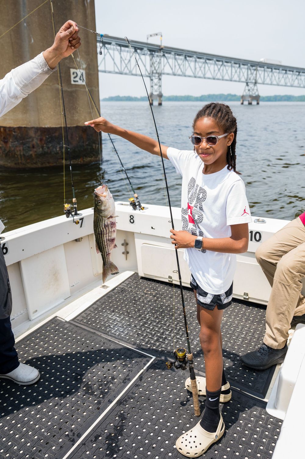 Young angler stands on a boat deck holding a fishing rod with a bridge visible in the background over the water.