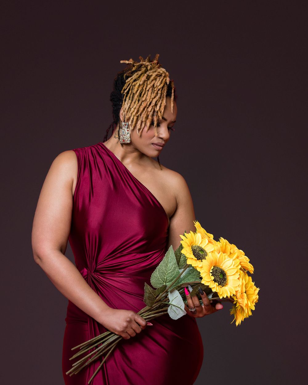 Figure in a burgundy one-shoulder dress holds a vibrant bouquet of yellow sunflowers against a dark background.