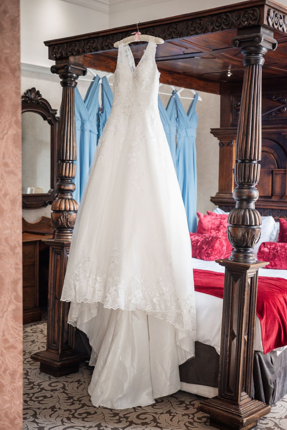 Elegant white lace wedding dress hanging on ornate wooden four-poster bed frame with blue bridesmaid dresses.