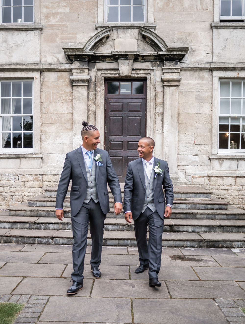 Two people in matching gray suits share a joyful moment walking together outside a historic stone building entrance.