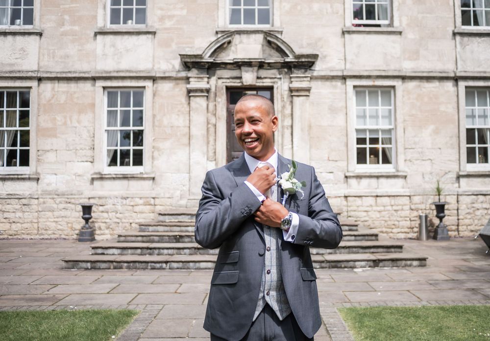 A person in a gray suit adjusts their tie while standing outside a grand stone building.