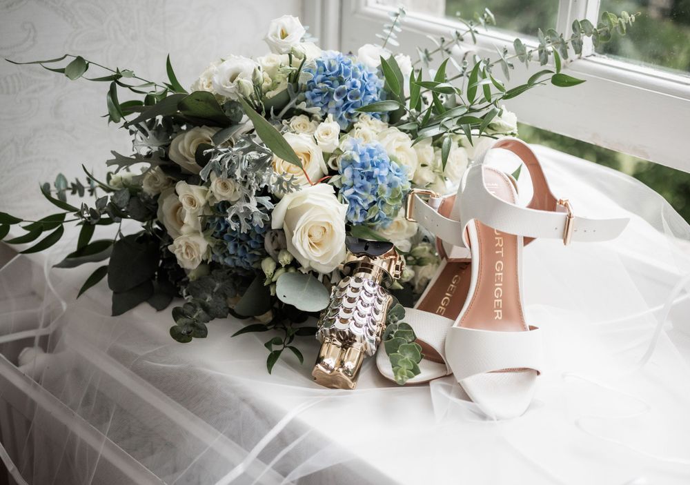 Elegant bridal bouquet with white roses and blue hydrangeas next to white wedding shoes on tulle fabric.