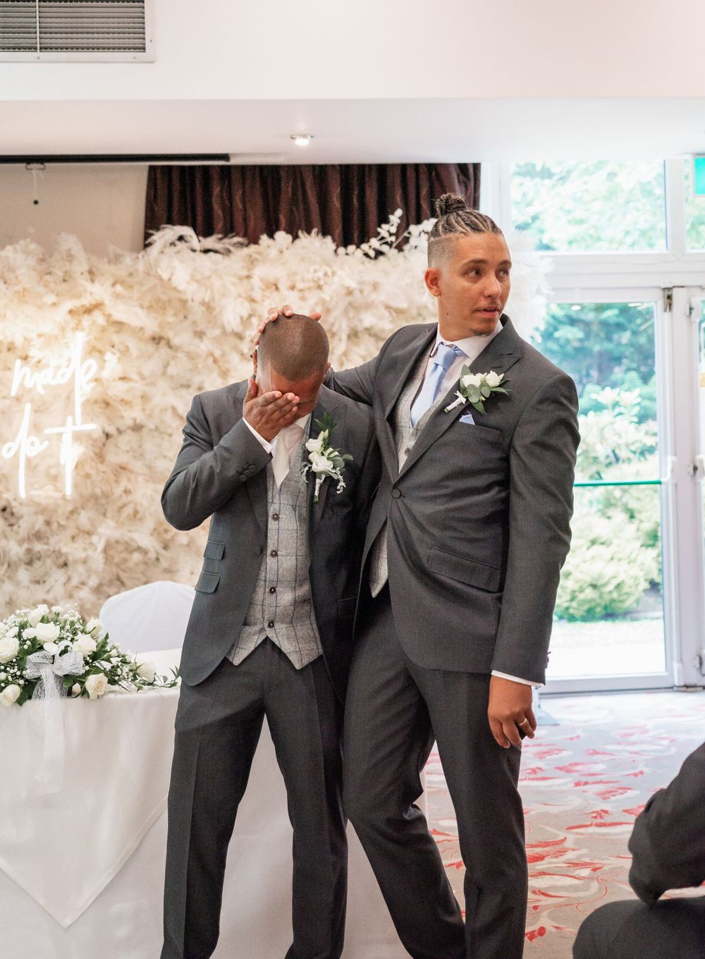 Two men in gray suits share an emotional moment at a wedding ceremony.