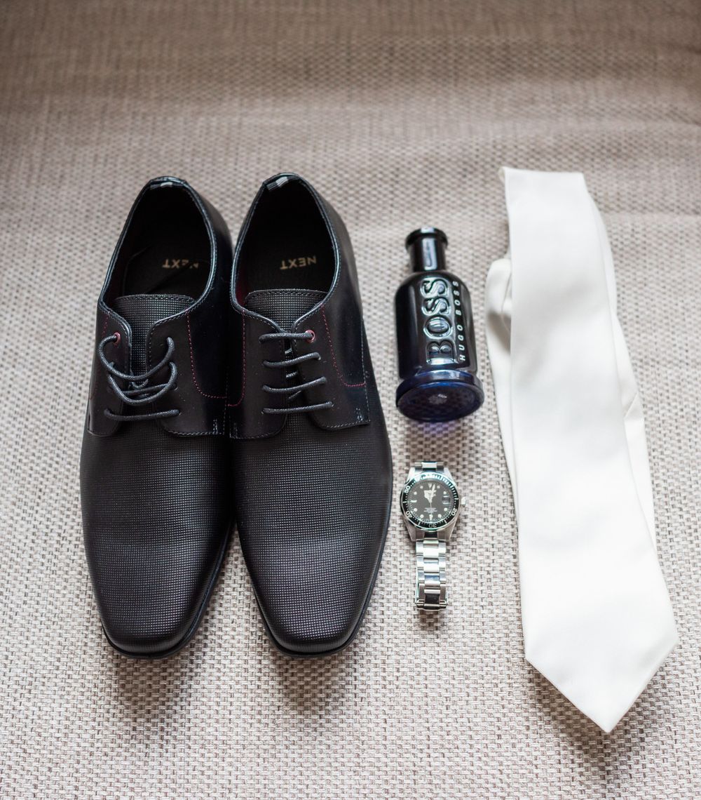 Groom's accessories featuring black dress shoes, cologne, watch, and white tie on beige fabric.