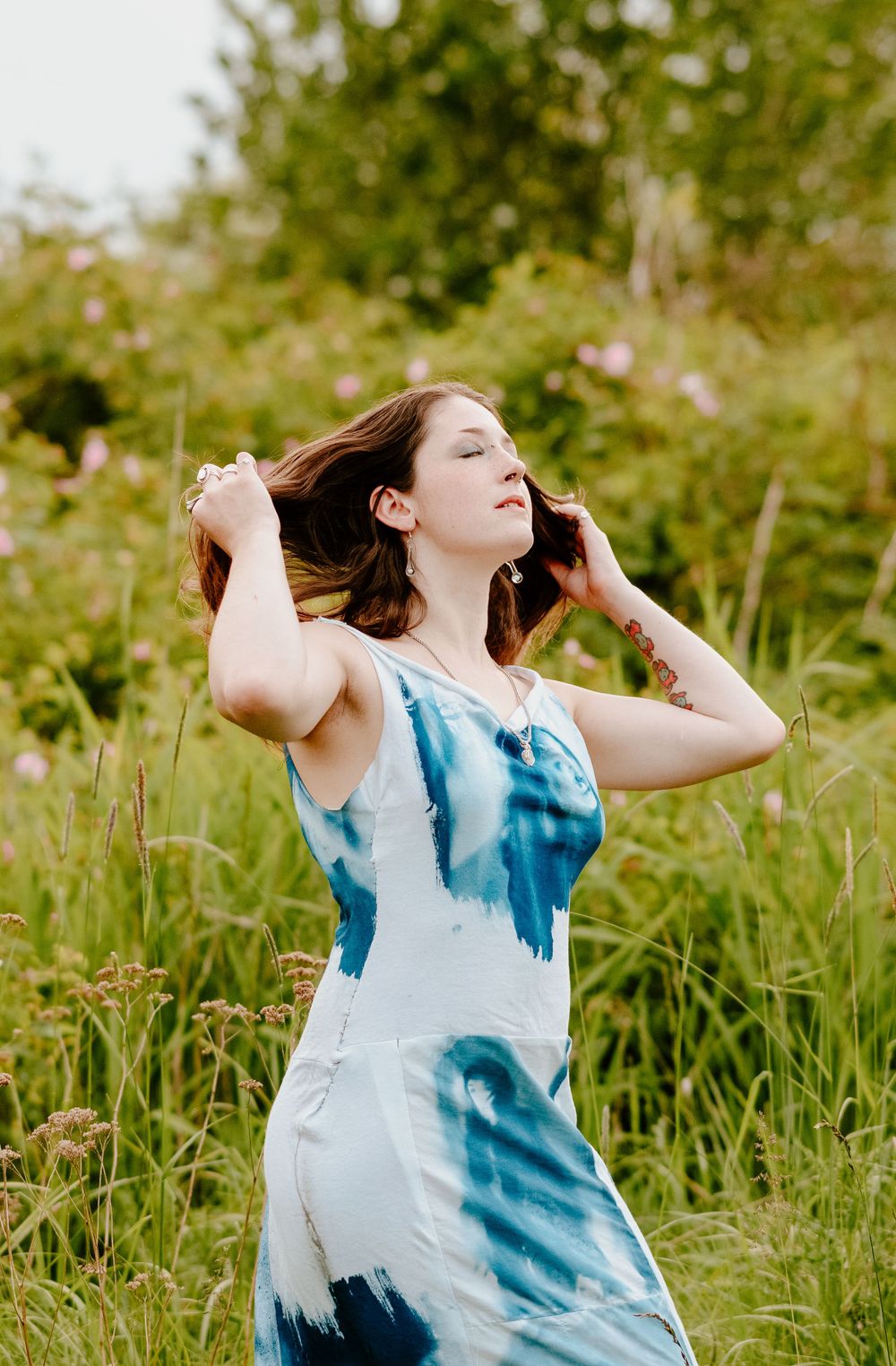 Woman in blue and white tie-dye dress poses playfully in a sunlit meadow with flowing hair.
