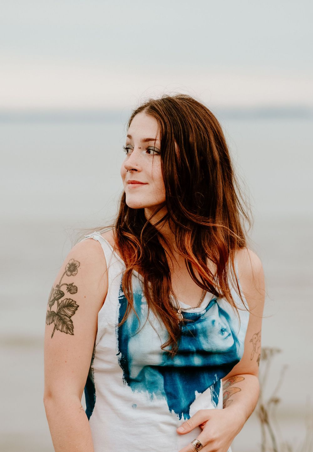 Woman in tie-dye dress with floral arm tattoo poses against blurred coastal background.