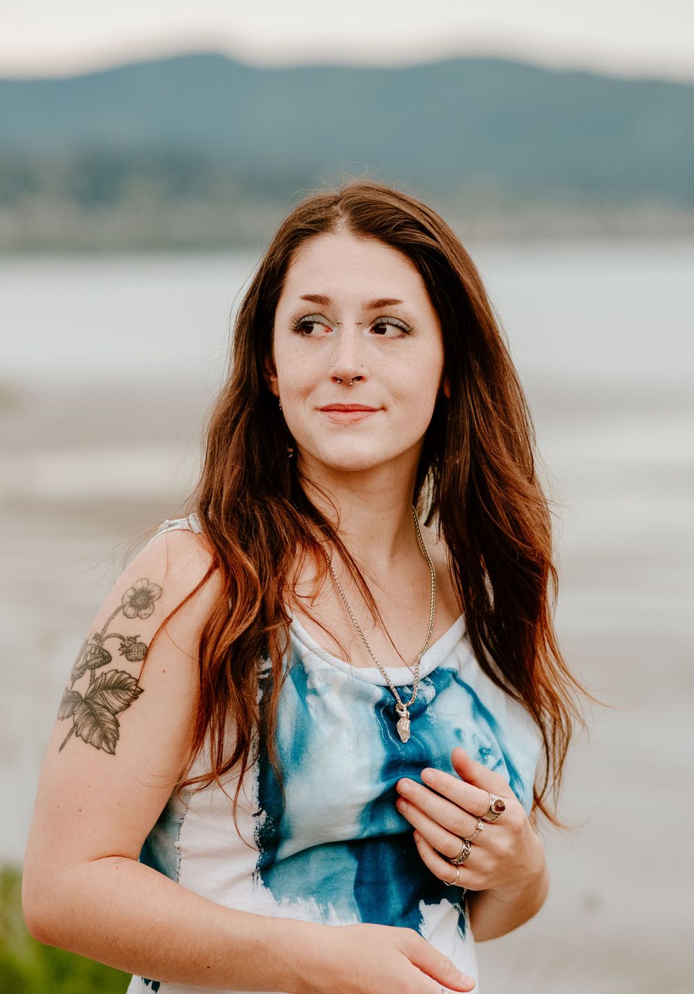 Person wearing blue tie-dye dress with tattoo stands near waterfront with mountains in background.