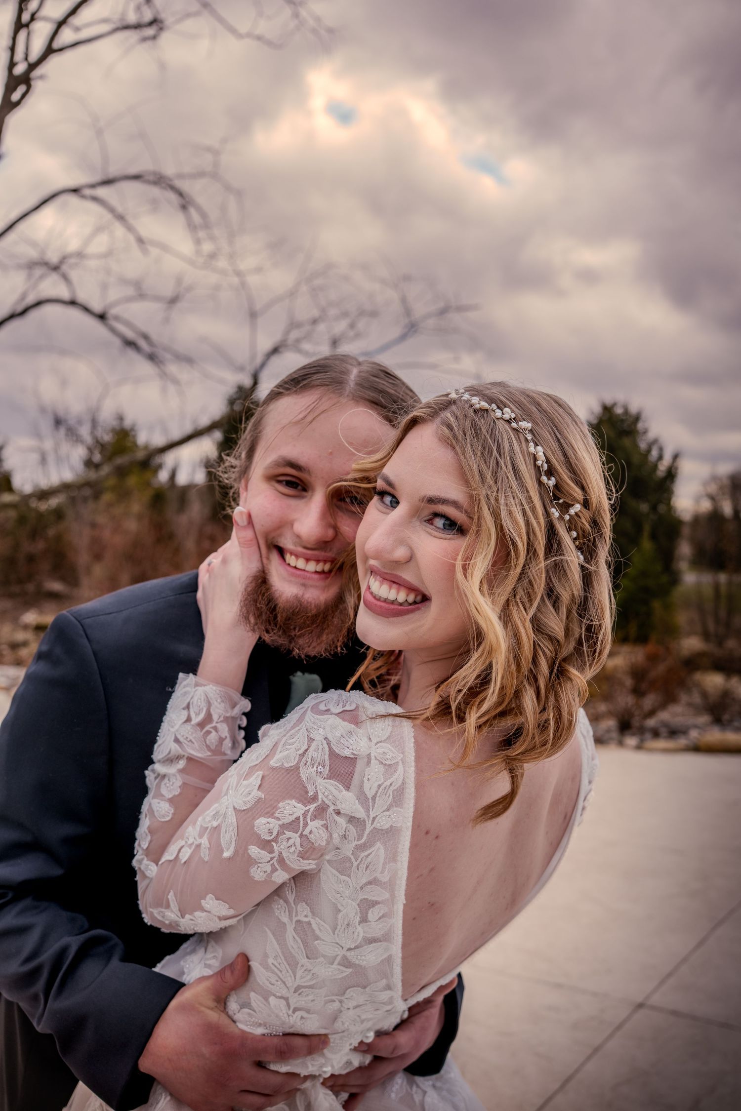 Smiling bride in lace gown with floral hair piece embraced by groom in navy suit outdoors under cloudy sky.
