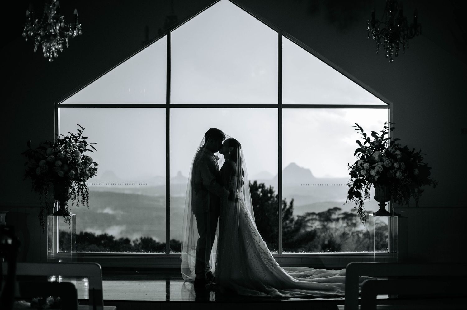 A romantic silhouette of a couple stands against a large triangular window with mountain views at sunset.