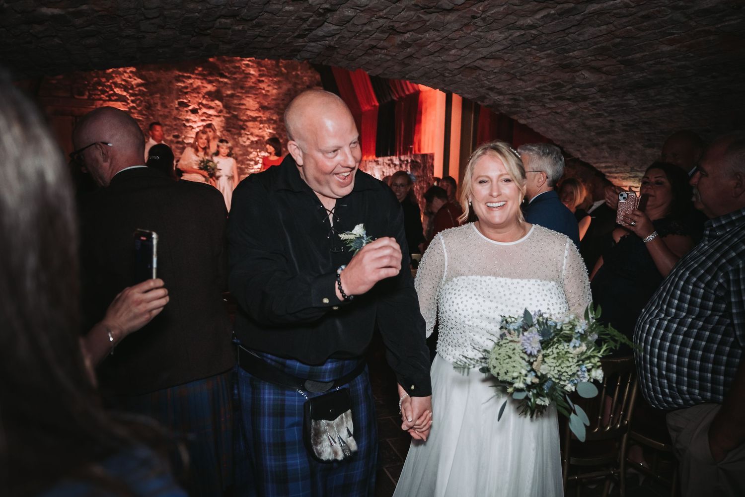 A couple celebrate at an indoor event in a stone-walled venue with moody red lighting and a bouquet of white flowers.