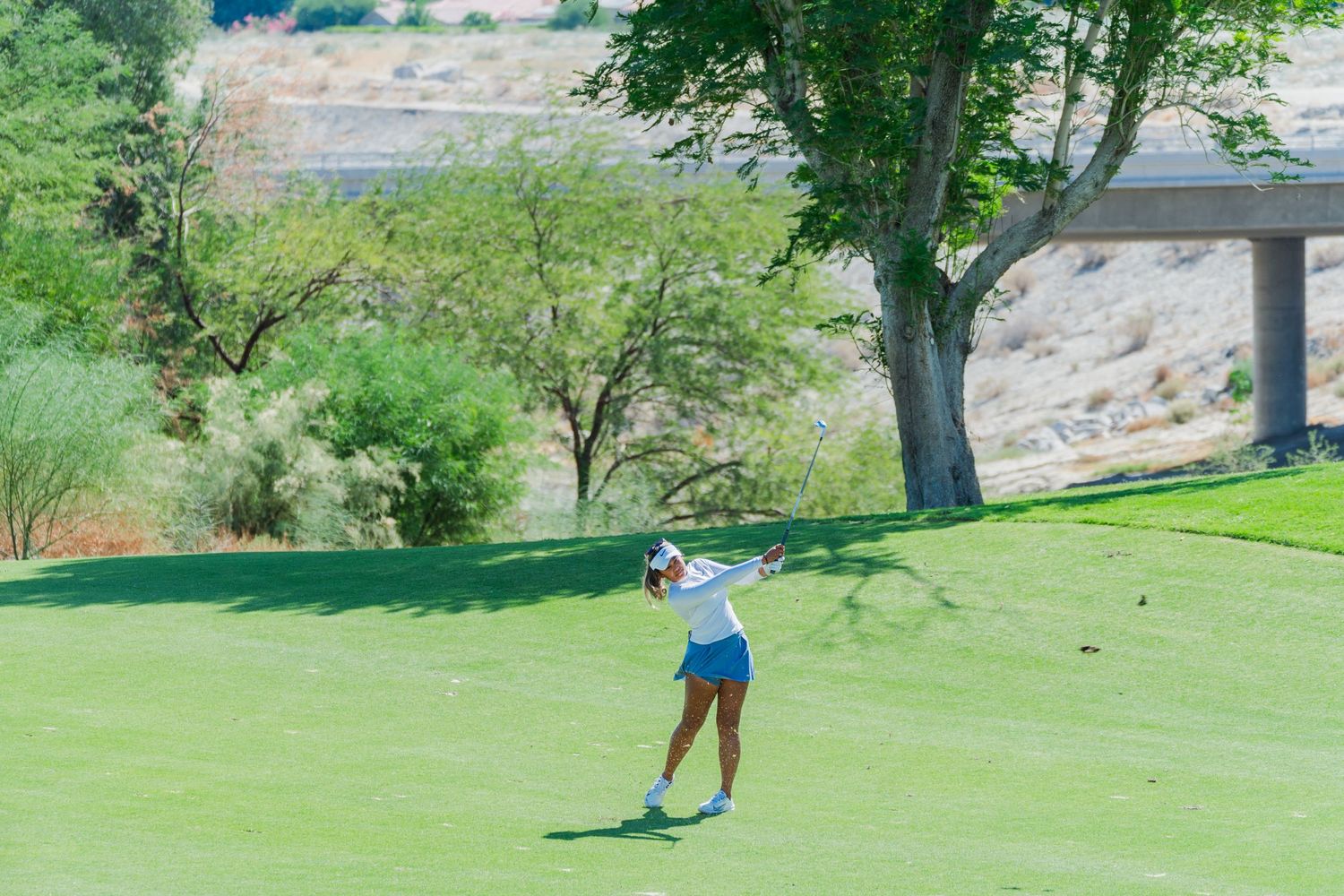 A golfer takes a swing on a lush green course with trees and hills in the background.