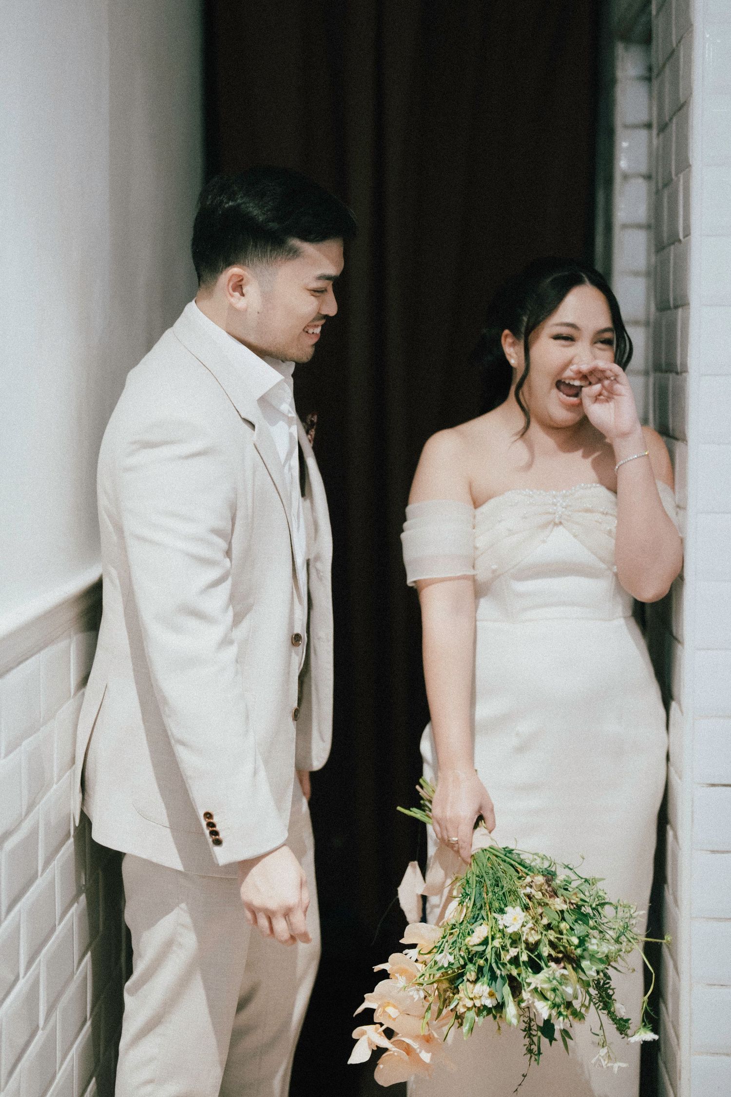 Bride in off-shoulder white gown laughing with groom in cream suit holding floral bouquet.