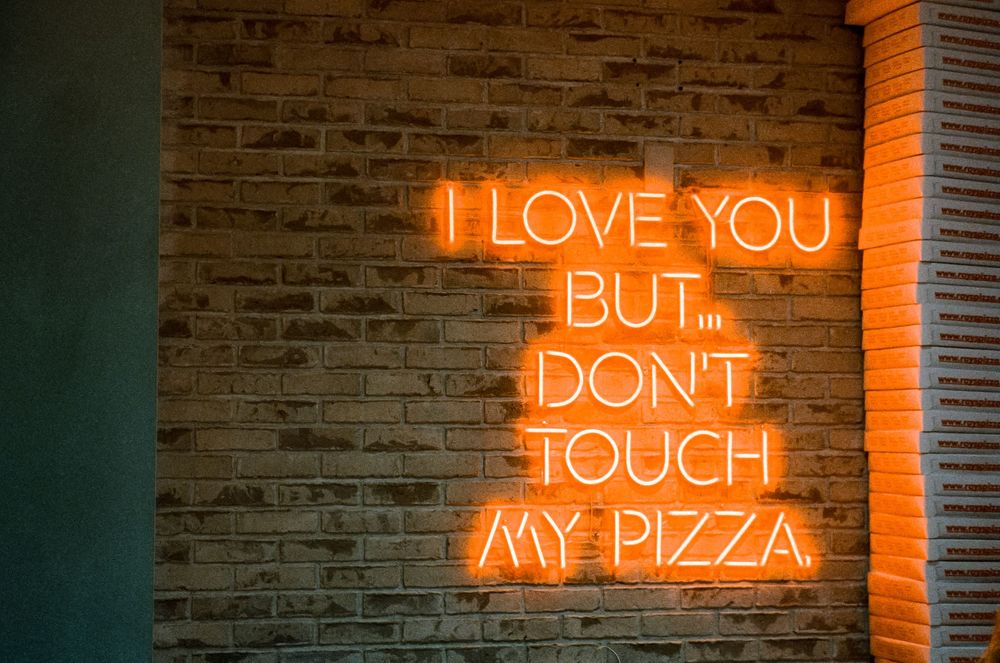 Orange neon sign on brick wall reads 'I love you but don't touch my pizza'.