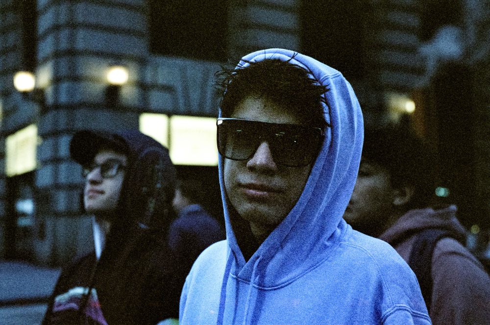 Street scene with person wearing blue hoodie and sunglasses at night.